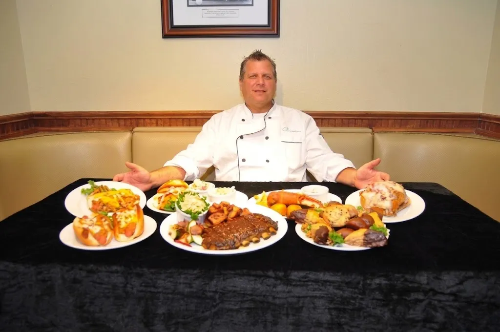 A Christo's chef at a booth behind a table spread with plates: coneys, ribs, sandwiches and desserts